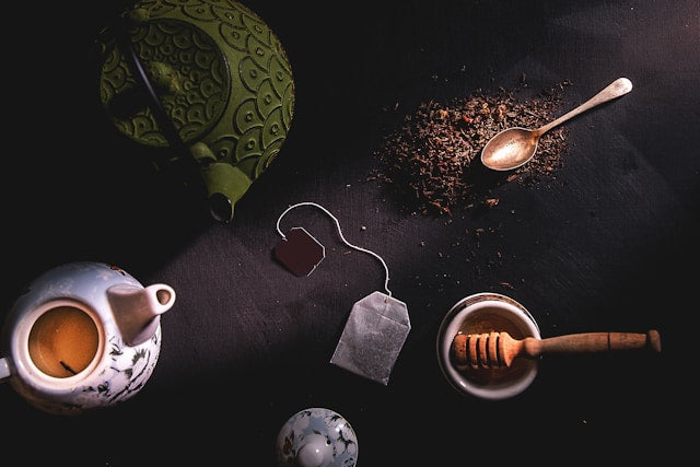 Tea-making items including a teapot, tea leaves, a spoon, a honey dipper, and tea bags on a dark surface.